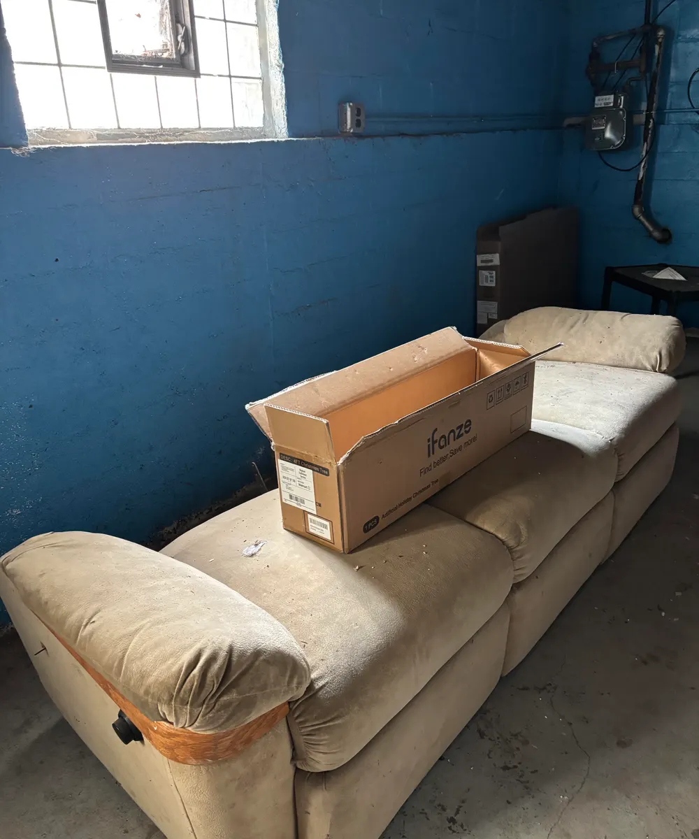 Worn beige reclining sofa with an empty cardboard box on it in a block-wall basement, ready for junk removal