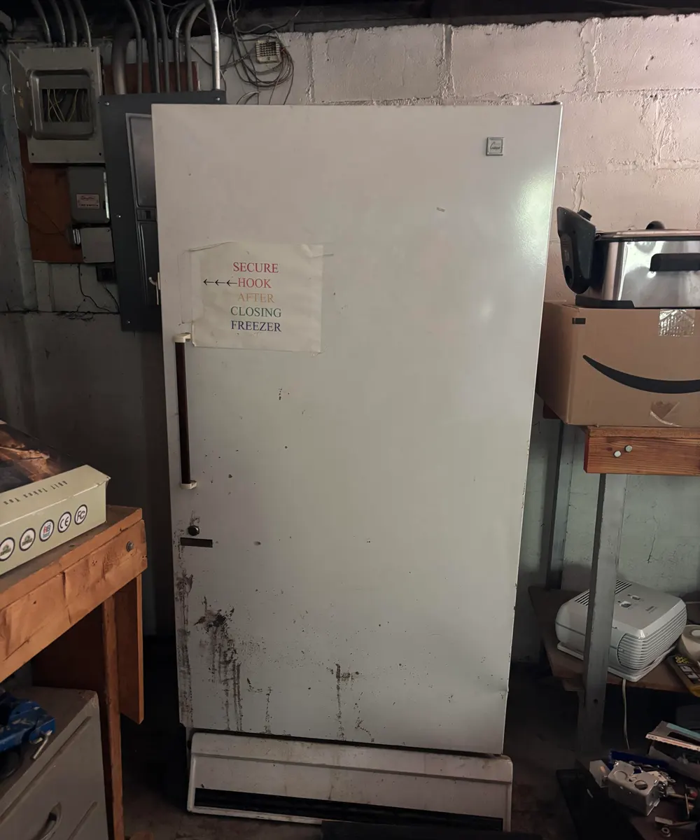 Old white upright freezer with a colorful 'secure hook after closing freezer' sign taped to the door in a Chicago basement