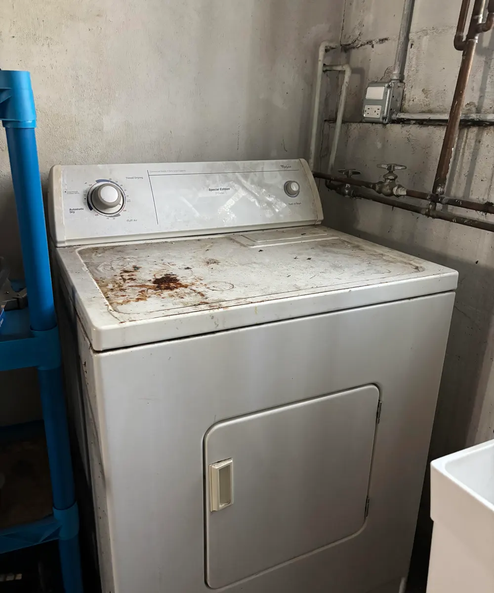 Old white Whirlpool electric dryer next to copper pipes in an unfinished basement laundry corner